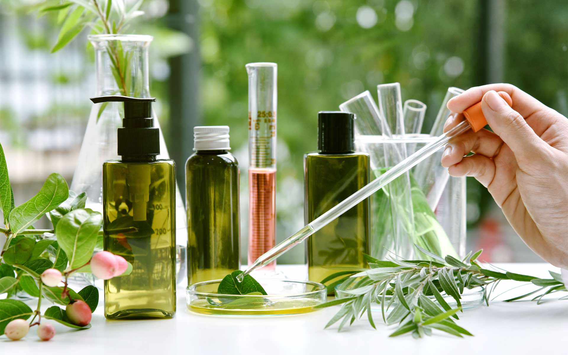 a hand holds a pipette dispensing essential oils amidst apothecary bottles and herbs on a white table 